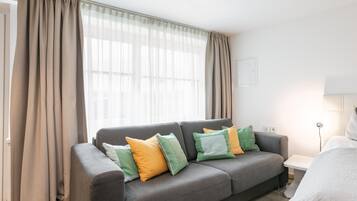 Een moderne woonkamer met een grijze bank, meerdere kussens, een lamp en een raam met dunne gordijnen.