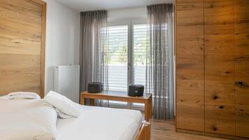 A bedroom with a bed, a wooden wardrobe, a radiator, a table with a bag, and a view through a window with curtains.