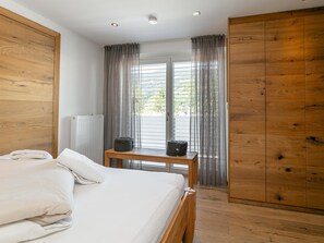 A bedroom with a bed, a wooden wardrobe, a radiator, a table with a bag, and a view through a window with curtains.