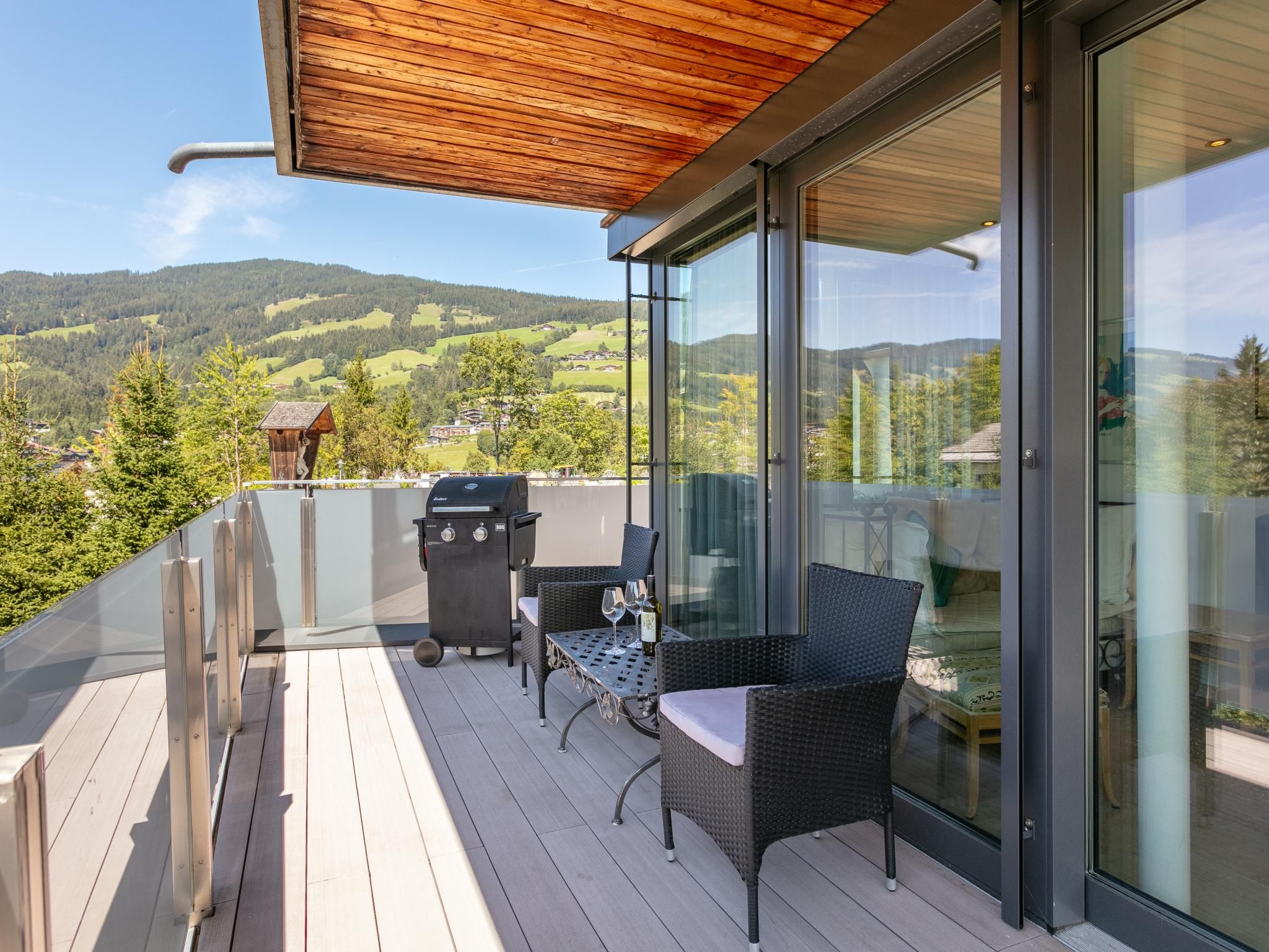A balcony with a wooden deck, a glass door, wicker furniture, and a barbecue grill.