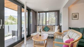 Un vaste espace de vie comprenant une table en bois, un canapé et un fauteuil. De grandes fenêtres offrent une vue sur l’extérieur.