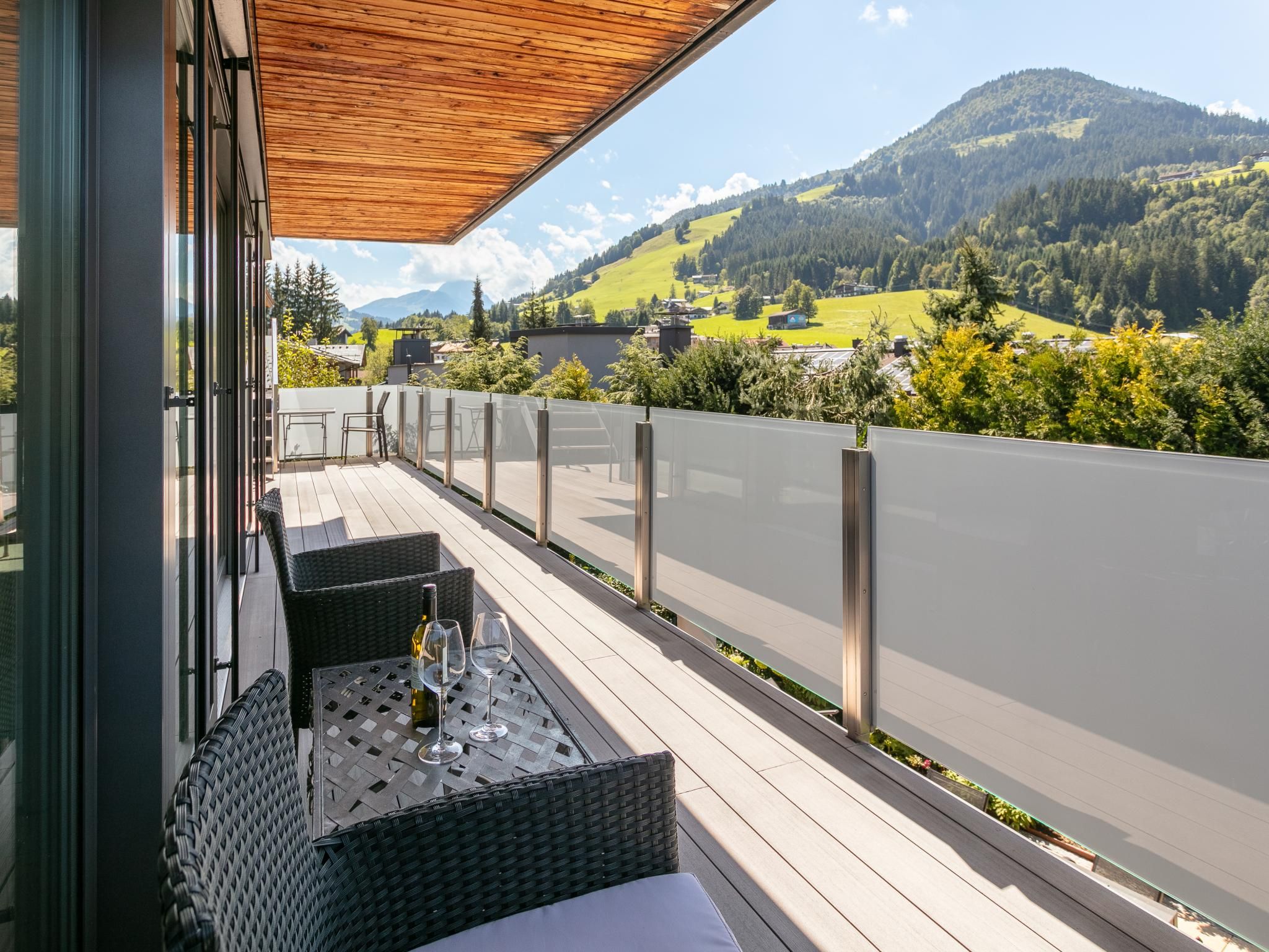 A terrace with wicker furniture, a bottle of wine, and glasses, overlooking a mountainous landscape.