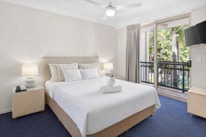 Une chambre d’hôtel comprenant un lit, deux tables de chevet, un ventilateur de plafond, une télévision et un balcon avec vue sur la verdure.