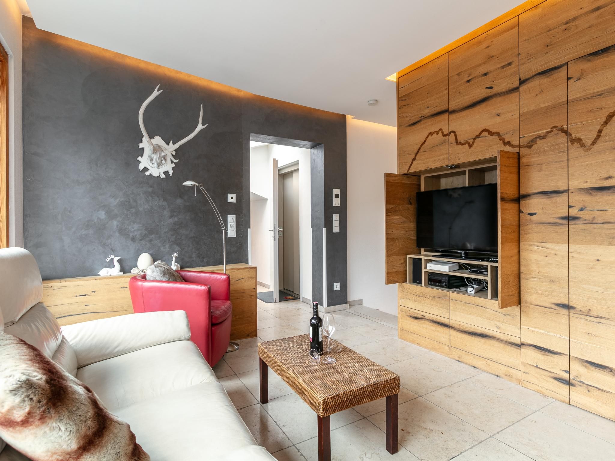 Una sala de estar moderna con un sofá, una butaca roja, una mesa de centro, un televisor montado en la pared y un panel de madera decorado con un cuerno de ciervo.