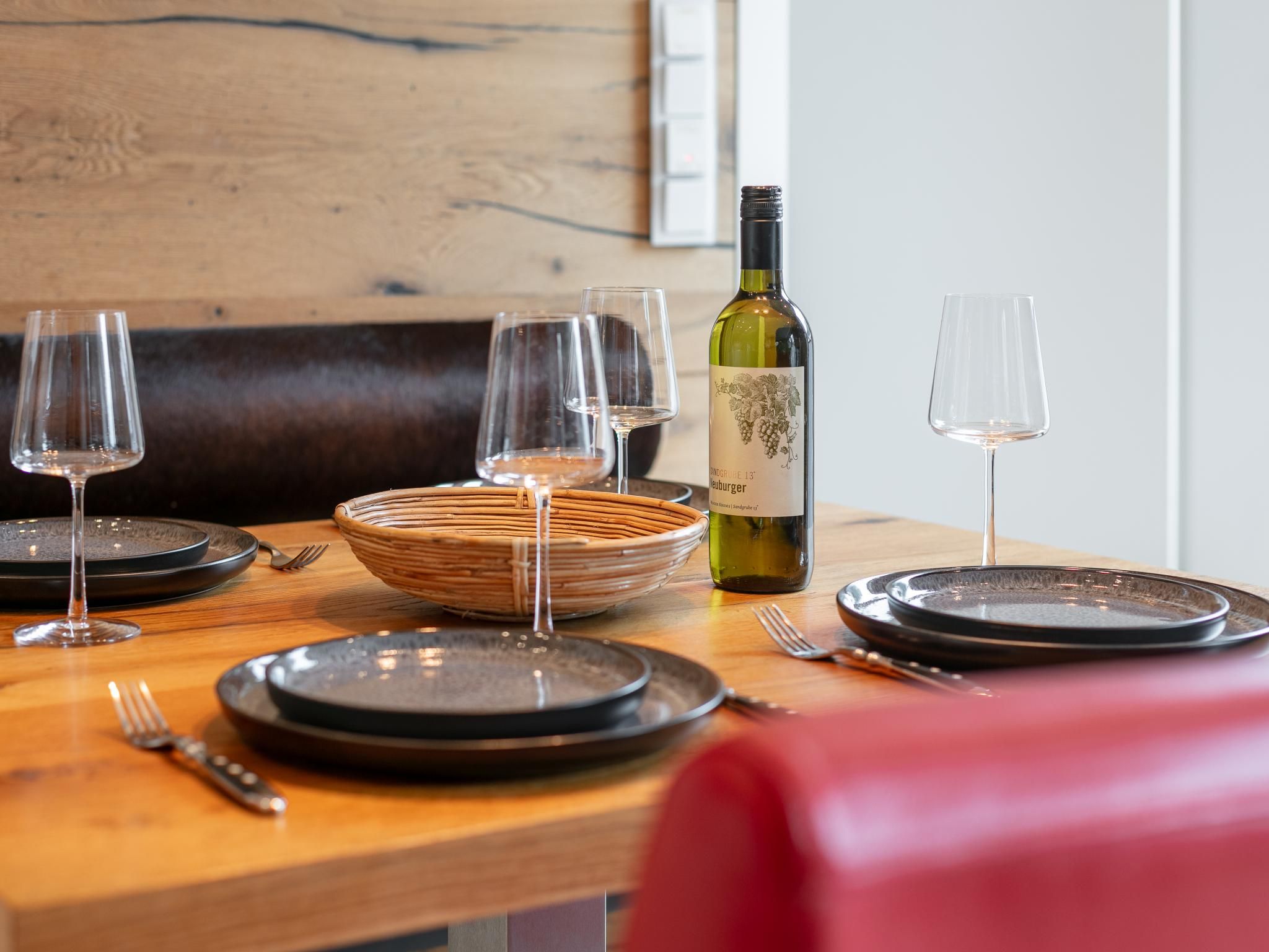 Une table à manger dressée avec des assiettes, des verres et une bouteille de vin, avec un panier en osier et une chaise rouge au premier plan.