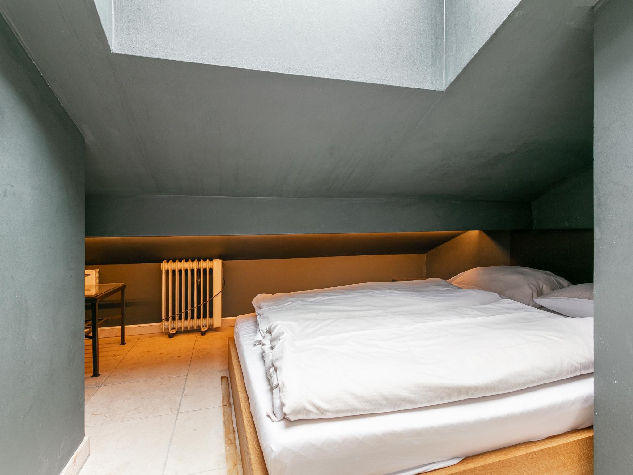 Um quarto de sótão pequeno e com pouca luz, com uma cama de solteiro, um radiador e uma mesa.