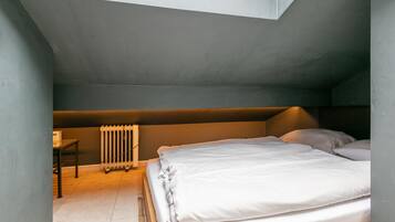 Um quarto de sótão pequeno e com pouca luz, com uma cama de solteiro, um radiador e uma mesa.