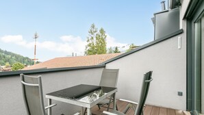 Un pequeño balcón exterior con una mesa y dos sillas, con vista a un tejado rojo y un páramo boscoso.