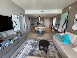 A modern living room with a sofa, a coffee table, a flat-screen TV, and a dining area with a table and chairs. - High Floor 2BR-2BA | Resort & Parking Fee Included (Hollywood)