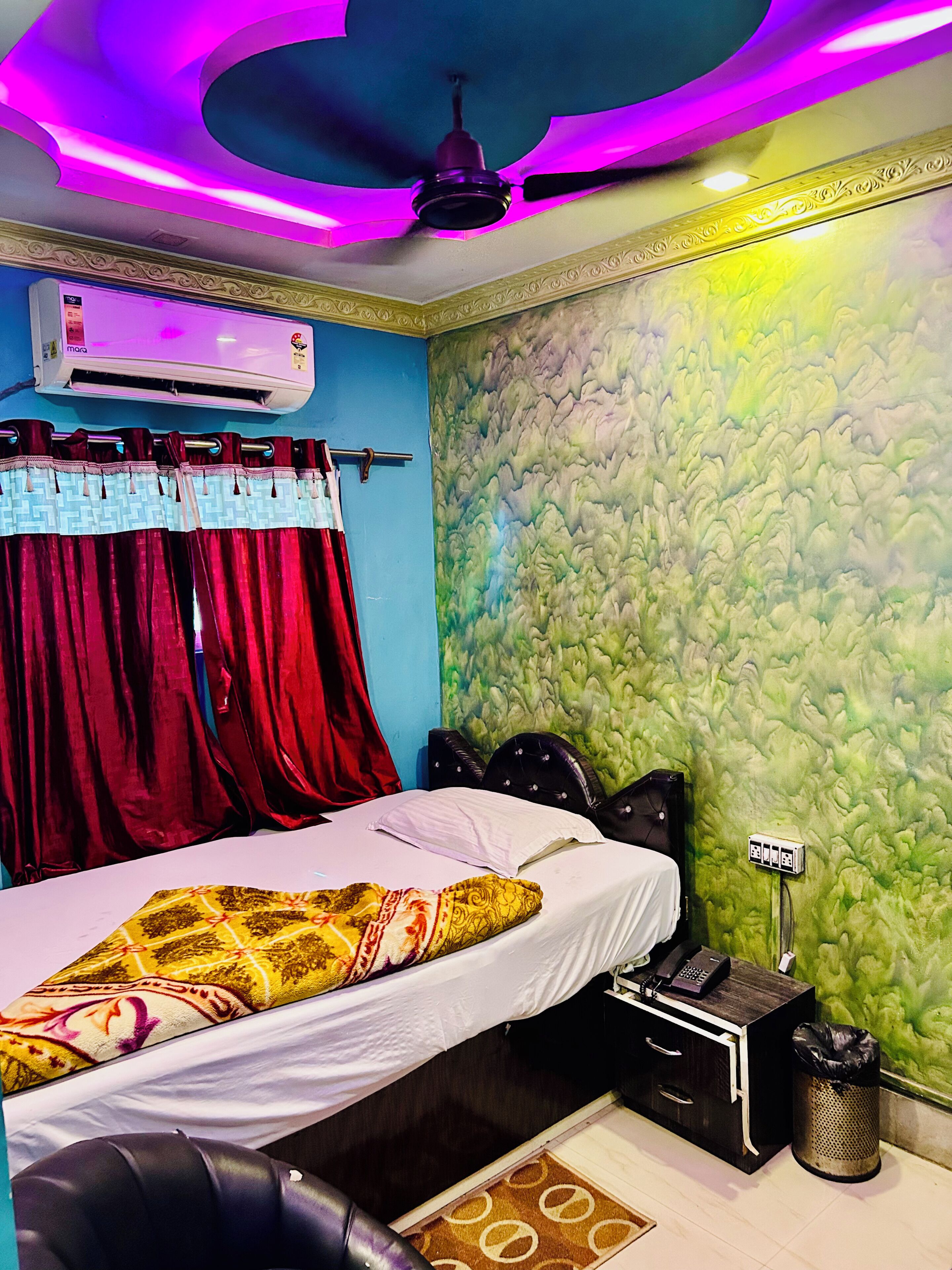 A bedroom with a bed, a ceiling fan, an air conditioning unit, a wall-mounted telephone, and a trash bin.