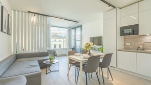 Un saló modern amb un sofà gris, una taula de dinar amb cadires, una taula auxiliar petita amb una planta, i una cuina amb armaris blancs i un microones.
