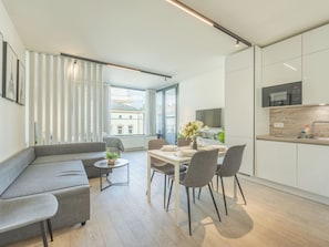 Ein modernes Wohnzimmer mit einem grauen Sofa, einem Esstisch mit Stühlen, einem kleinen Couchtisch mit Pflanze und einer Küchenzeile mit weißen Schränken und einer Mikrowelle.