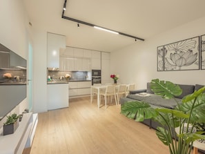A modern kitchen with white cabinets, a dining area with a white table and chairs, and a grey sofa in a living room with large green plants. - Nový Smíchov by Interhome (Smichov Kosire Jinonice)