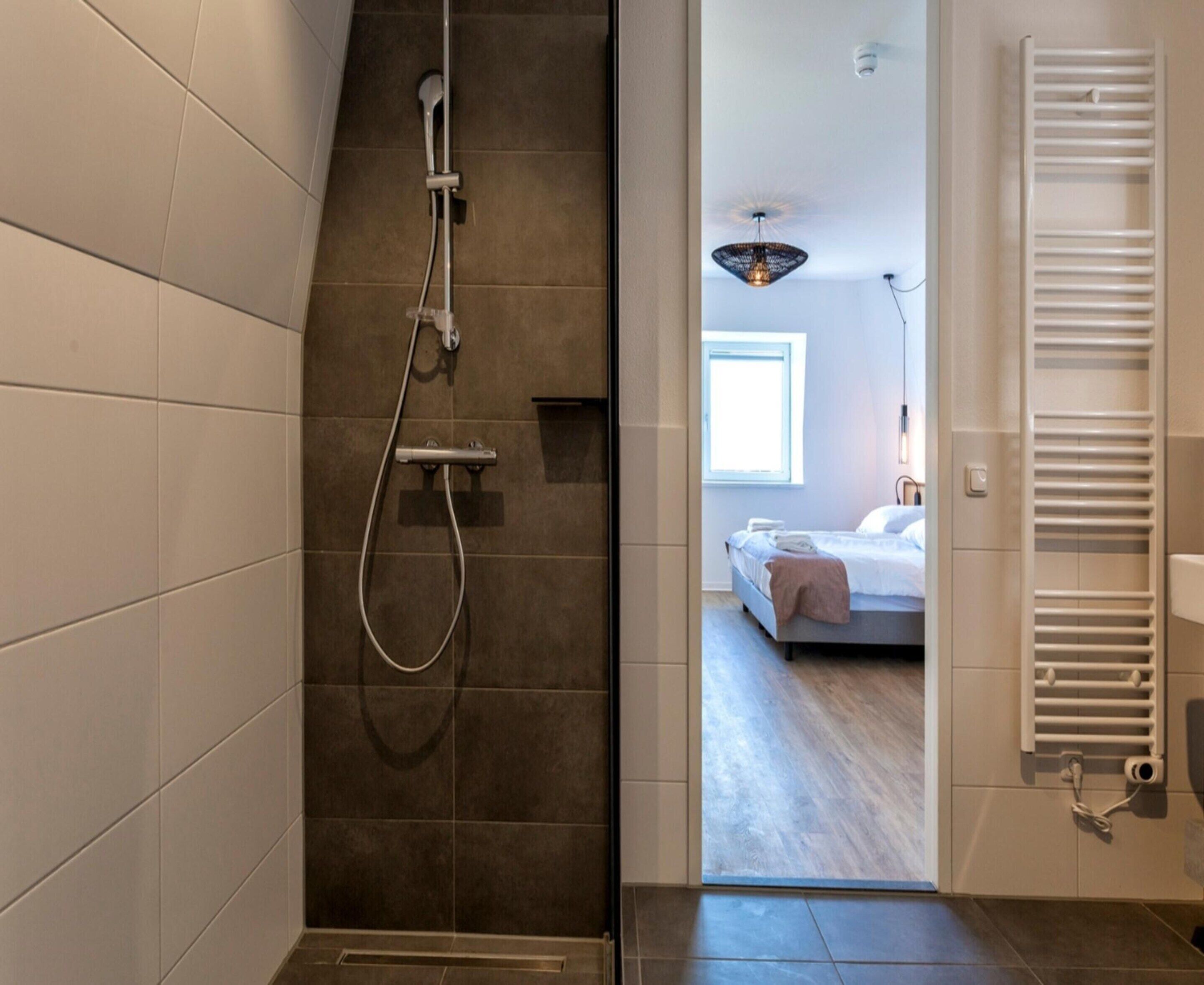 A modern bathroom with a shower, a visible towel rack, and a view into a bedroom with a bed and a window.