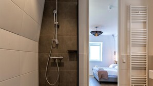 Une salle de bain moderne avec une douche, un porte-serviettes apparent et une vue sur une chambre à coucher avec un lit et une fenêtre.