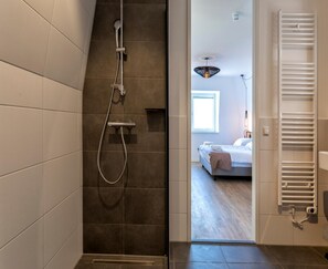 Ein modernes Badezimmer mit Dusche, einem sichtbaren Handtuchhalter und Blick in ein Schlafzimmer mit Bett und Fenster.