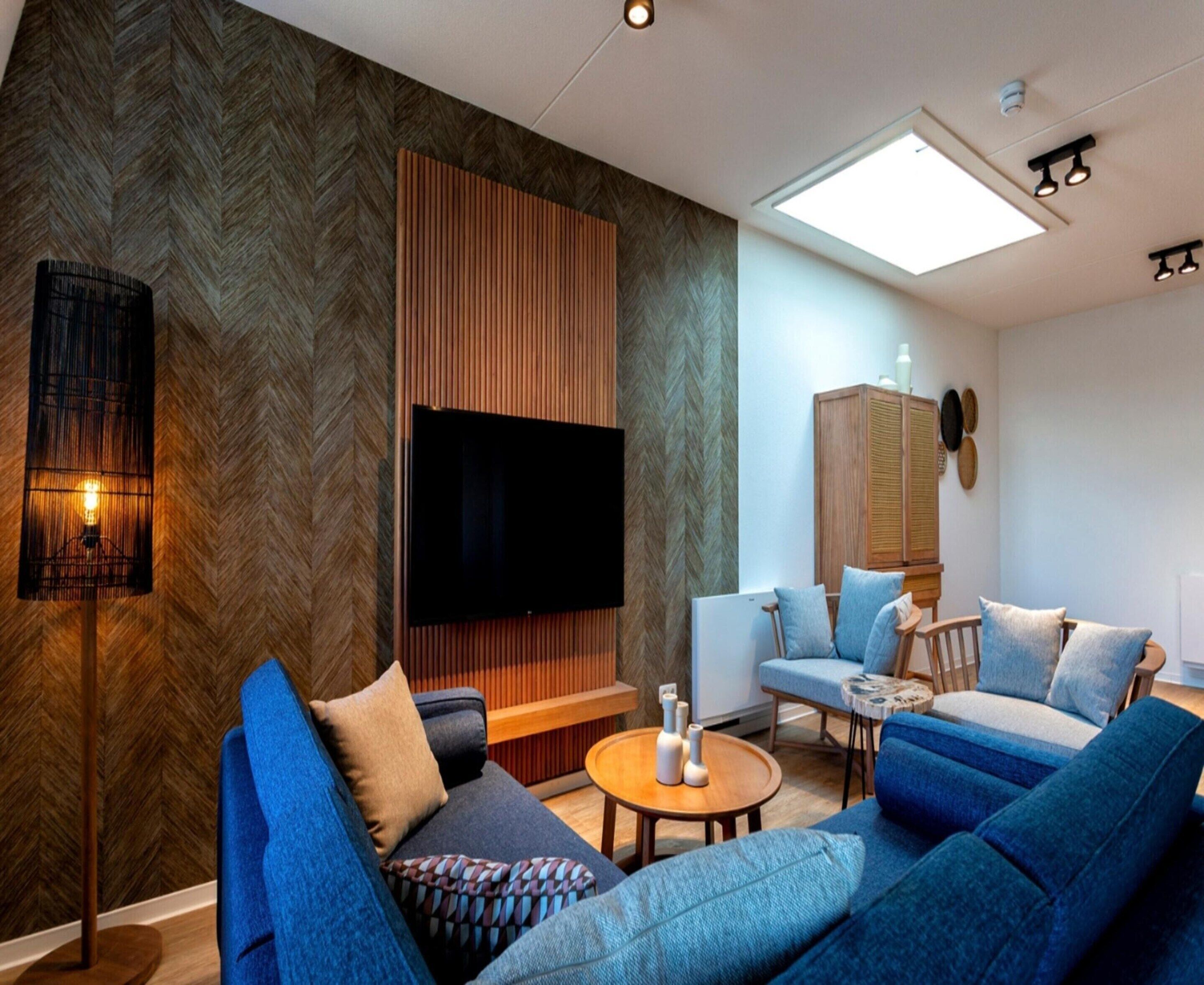 A modern living room with a large flat-screen TV, blue sofas, a wooden accent wall, and a ceiling light fixture.