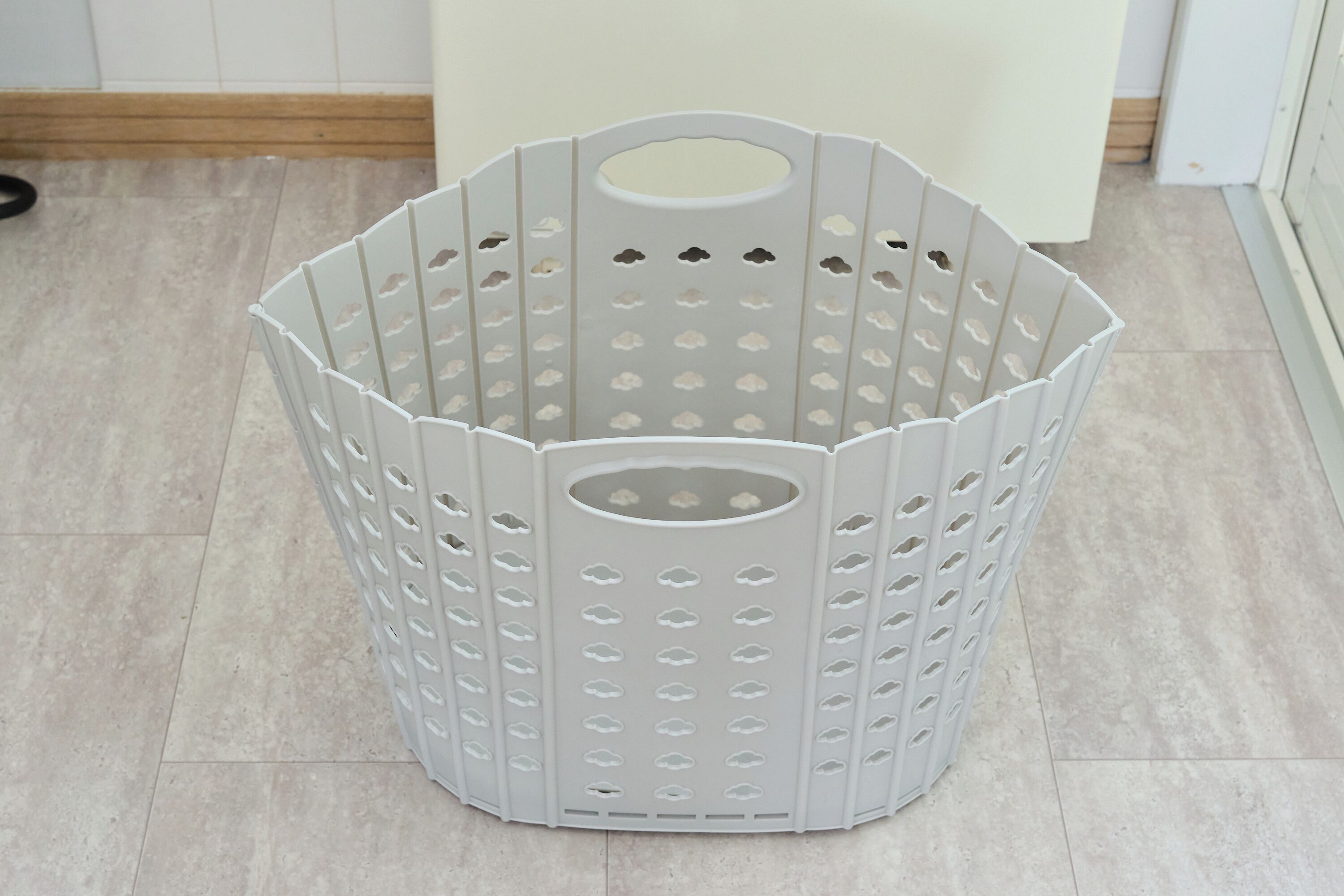 A grey, hexagonal-shaped laundry basket with multiple holes and a handle, placed on a tiled floor.
