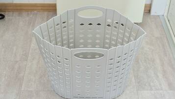 A grey, hexagonal-shaped laundry basket with multiple holes and a handle, placed on a tiled floor.