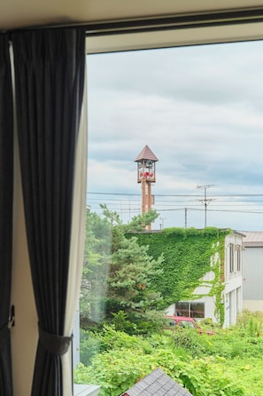 A view through a window with a curtain, featuring a tower with a red roof and a bell, surrounded by greenery and a red car.