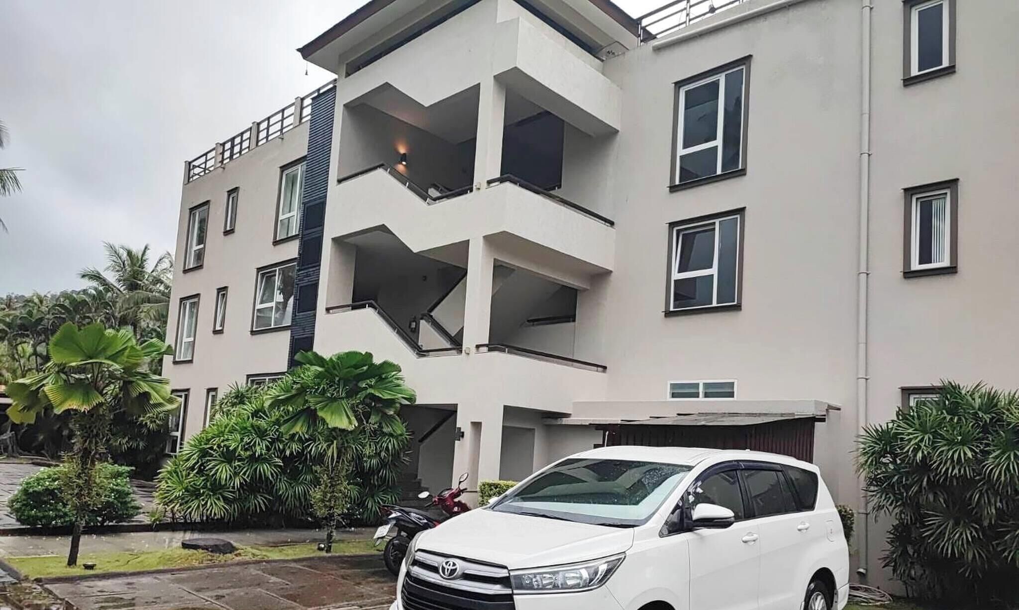 A multi-story building with balconies, a white car parked in front, and surrounding greenery.