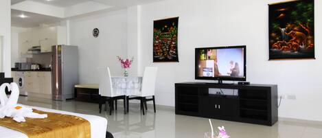 Habitación de hotel con cama, una mesa de comedor con sillas, televisor, refrigerador y una mesa de vidrio con una computadora portátil y una bebida.