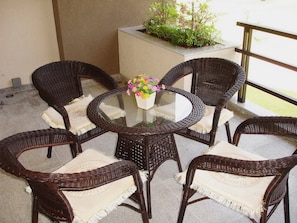 A round glass table surrounded by wicker chairs with cushions, a potted plant with flowers, and a concrete planter on a balcony.