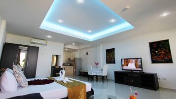 A modern hotel room with a bed, a dining area, a TV, and a kitchen in the background.