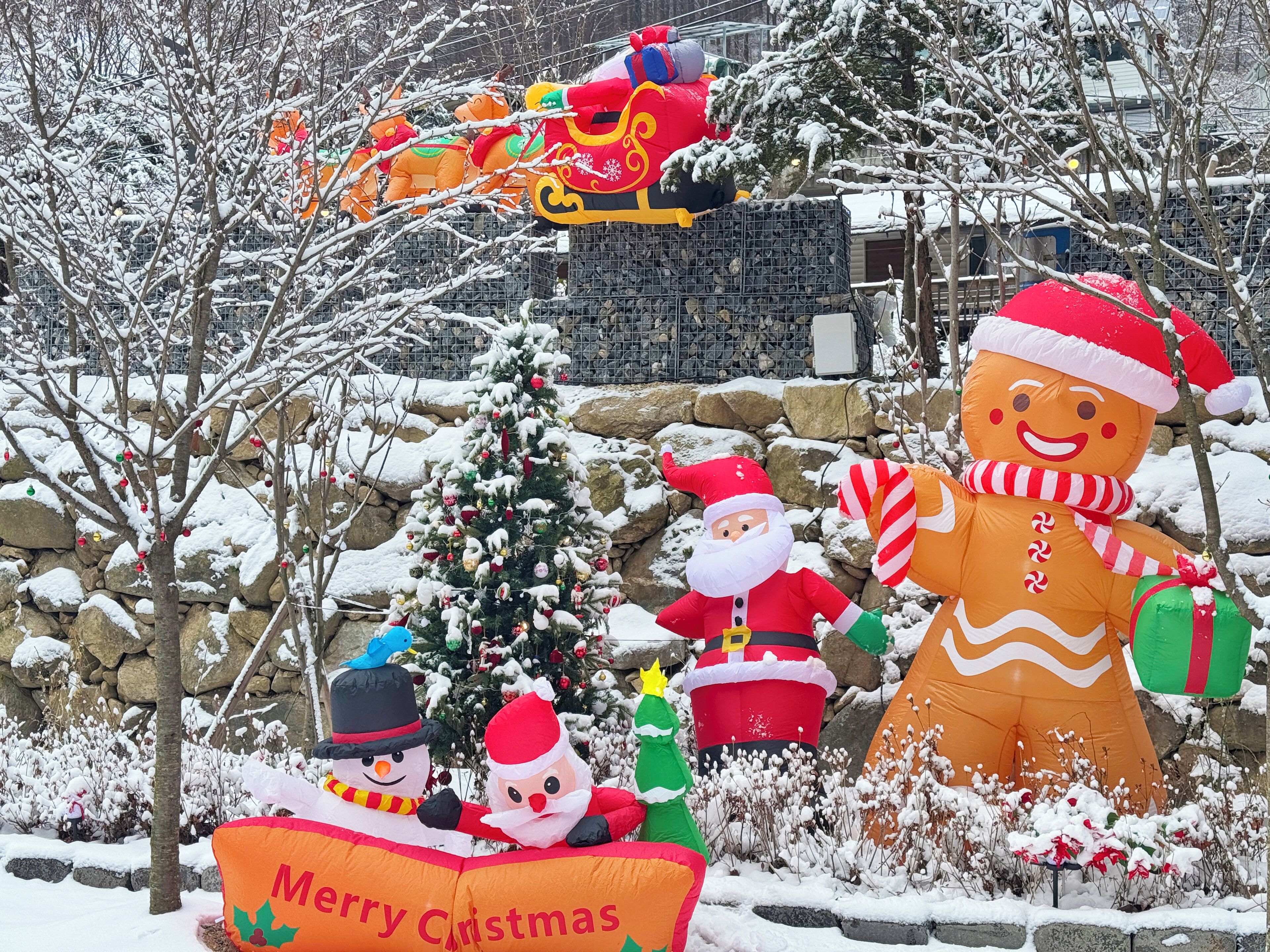 A snow-covered outdoor scene with a large inflatable gingerbread man, a snowman, and a Santa Claus figure.