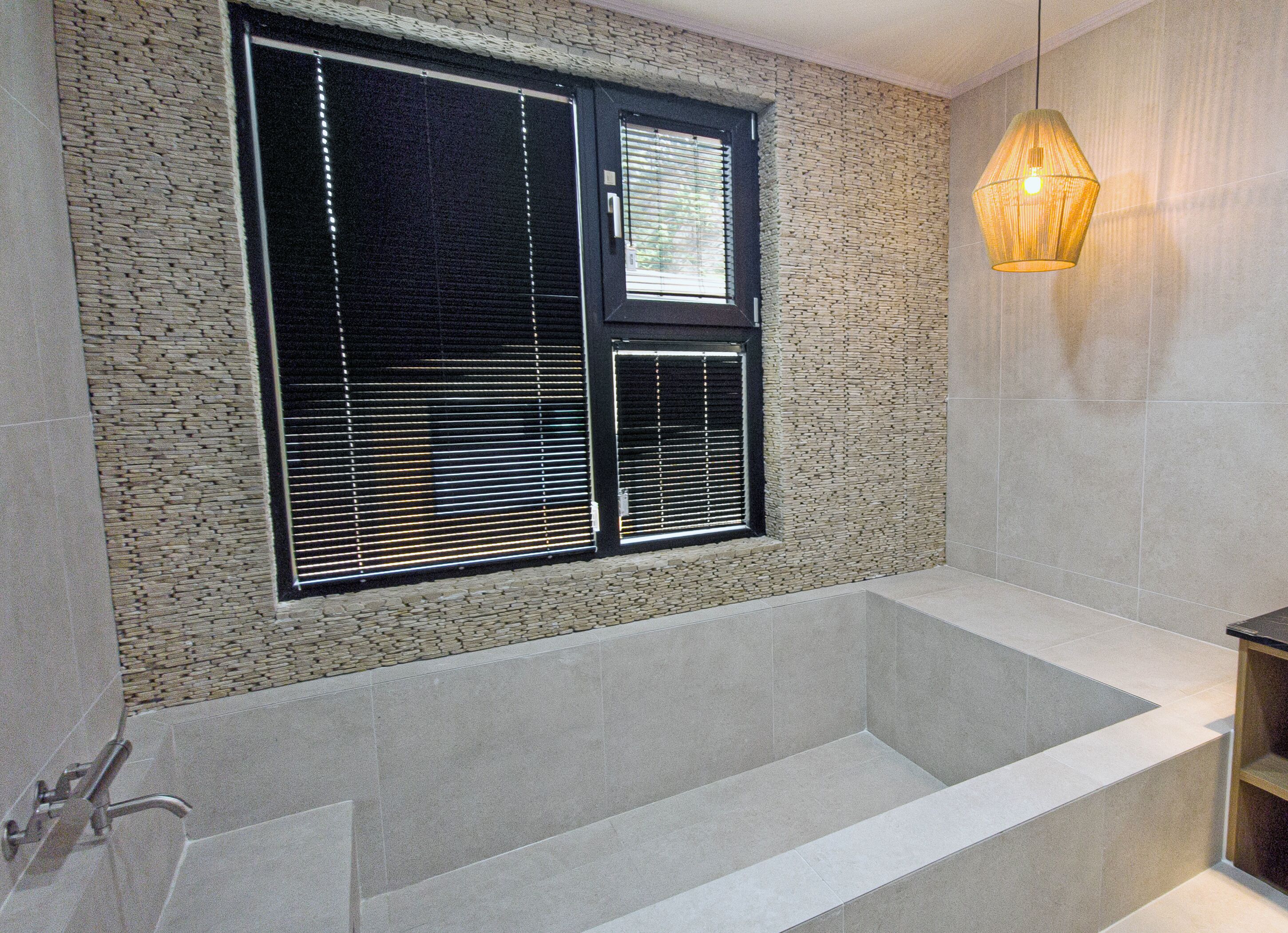 A modern bathroom with a large bathtub, a window with blinds, and a hanging light fixture.