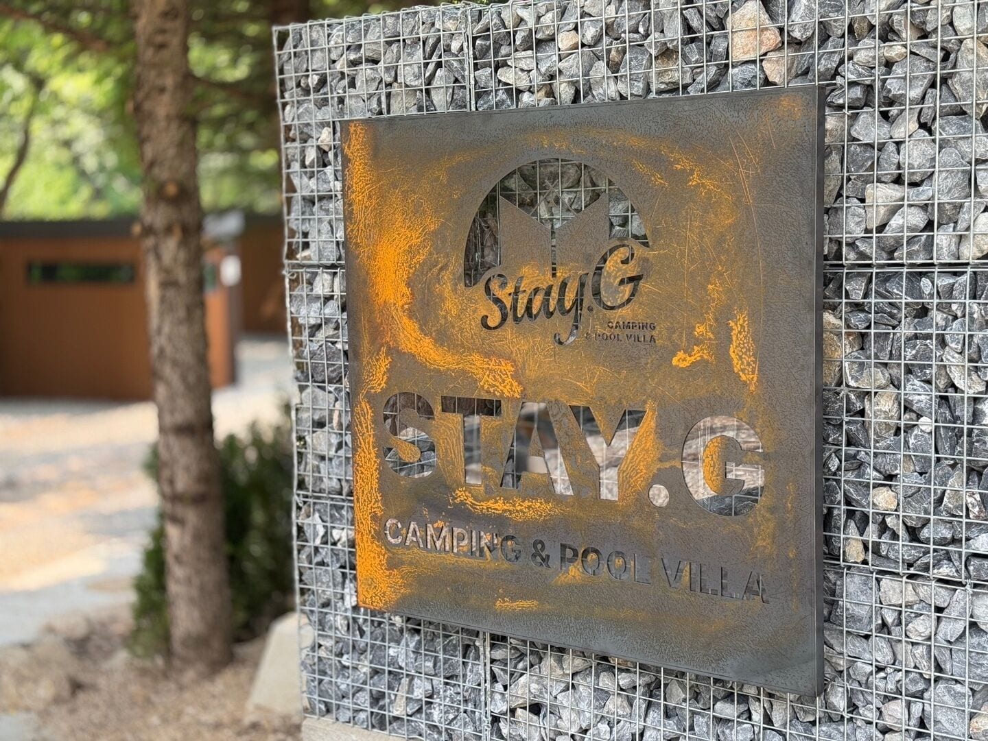 A metal sign with the Stay G logo and text 'STAY.G CAMPING & POOL VILL' mounted on a rock wall.
