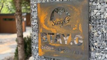 A metal sign with the Stay G logo and text 'STAY.G CAMPING & POOL VILL' mounted on a rock wall.