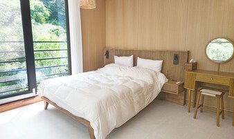 A bedroom with a large bed, a wooden headboard, a bedside table, a mirror, and a view of greenery outside.