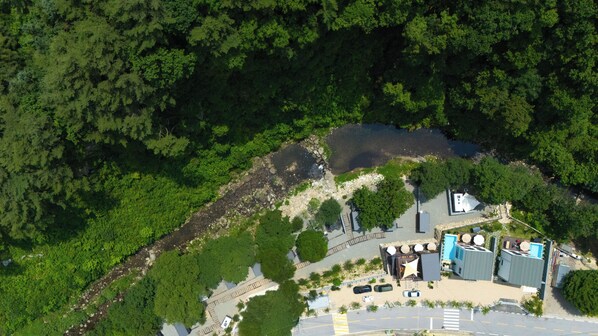 Vue aérienne d’une propriété comprenant une piscine, un petit bâtiment et un espace de stationnement, le tout entouré d’une forêt dense.