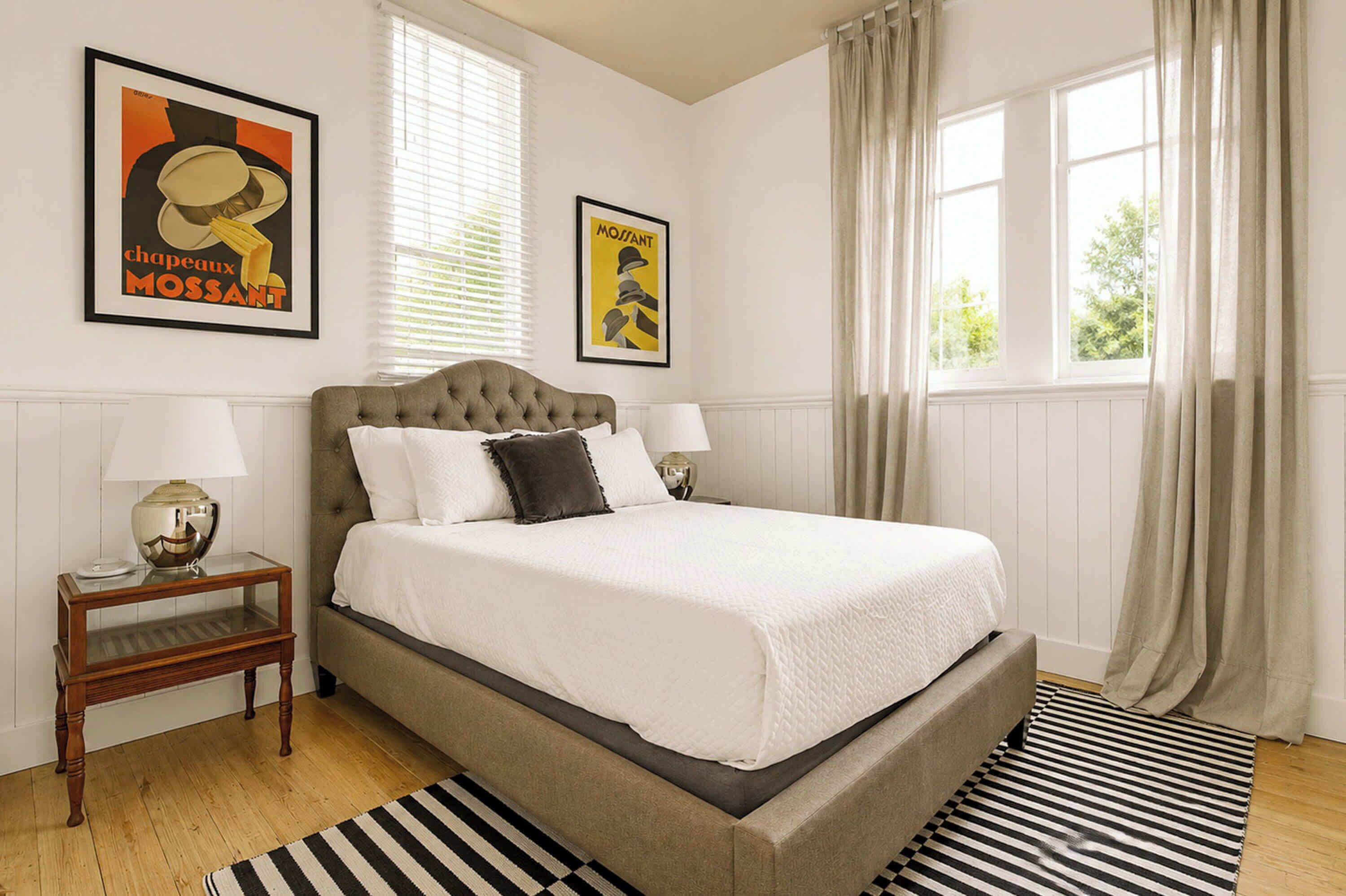 A bedroom with a large bed, two bedside tables with lamps, a striped rug, and two posters on the wall.