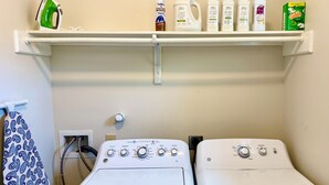 A laundry area with two front-loading washers, a shelf with various cleaning products, and a patterned towel hanging on a rack. - CasaDeCarrion: 30+Day Extended Corporate Stay Fully Furnished 3bd/2.5ba (San Antonio)