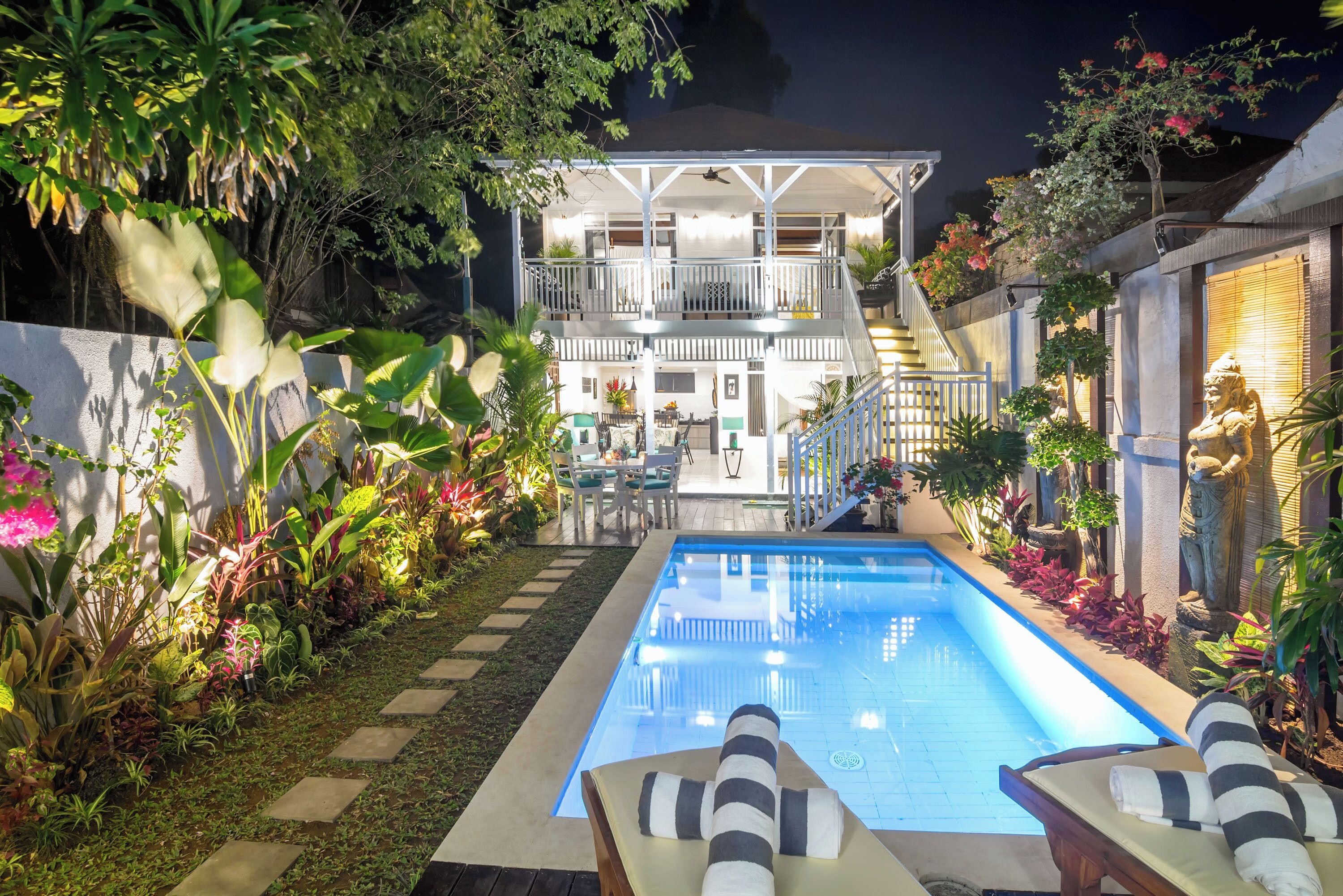 A swimming pool with lounge chairs, surrounded by lush greenery and a two-story building with a balcony.