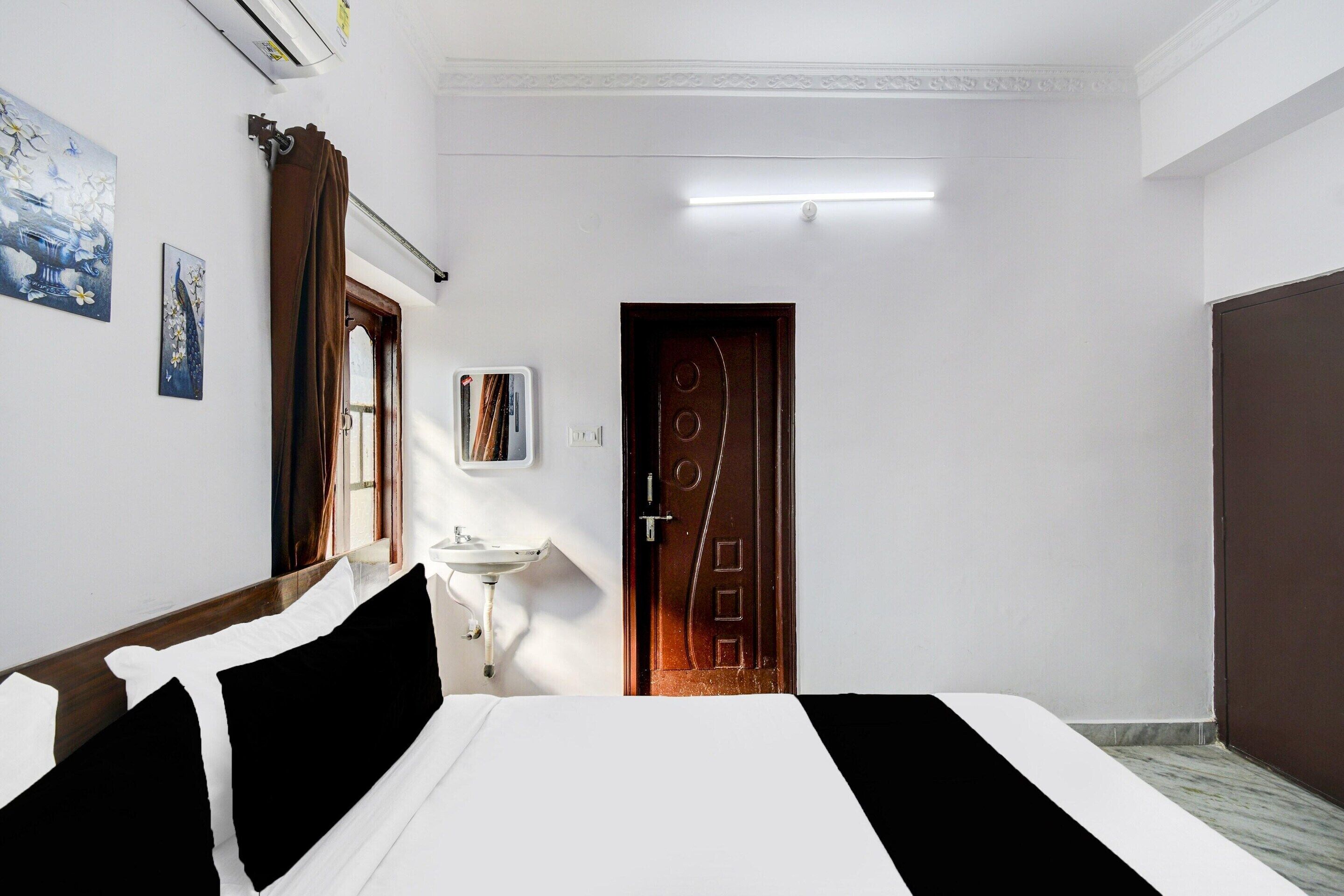 A hotel room with a bed, a bedside table, a mirror, a window with curtains, a wall-mounted air conditioner, and a door.
