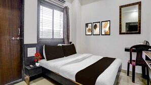 Habitación de hotel con cama, escritorio, silla, equipo de aire acondicionado, ventana con cortinas y obras de arte en la pared.