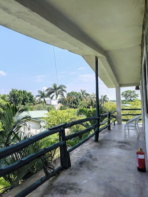 A covered outdoor area with a concrete floor, a metal railing, and a view of greenery and buildings in the distance. - Corbett Soho Resort (Kaladhungi)