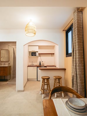 A compact kitchen with a microwave, a sink, and a window with curtains. There are two wooden stools and a table with plates. - Pointe au Sel Apartment - L'Escale Bleue, Saint-Leu, Réunion (Saint-Leu)