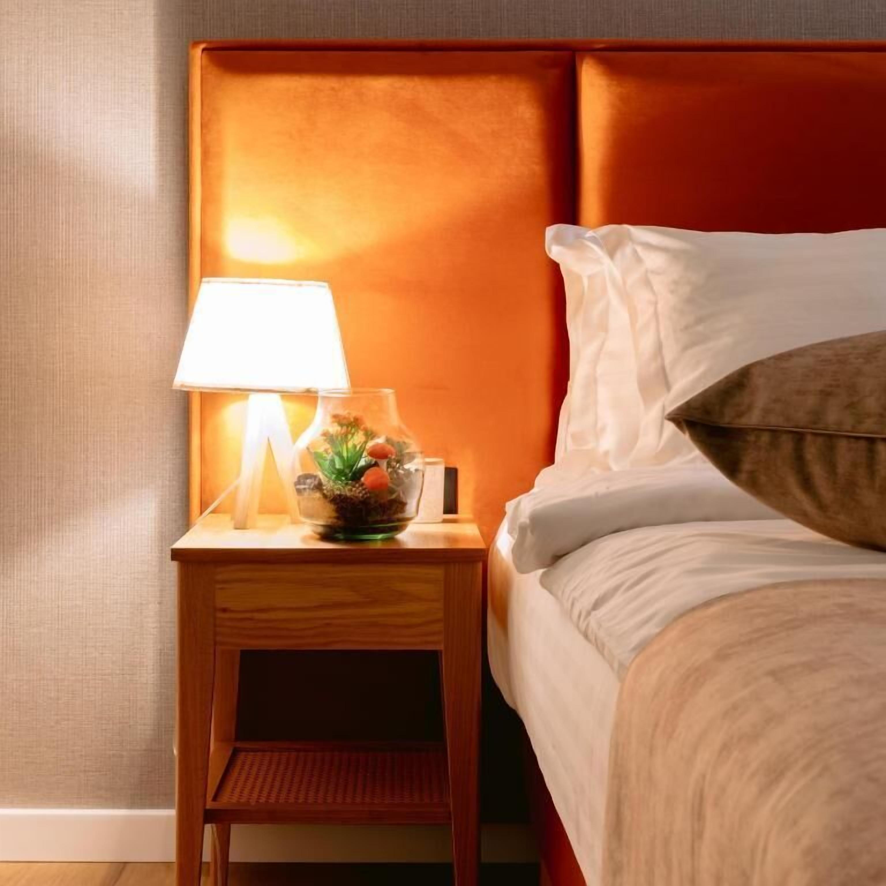 A neatly made bed with white linens and a brown pillow, a bedside table with a lamp and a small plant, and a wooden nightstand.