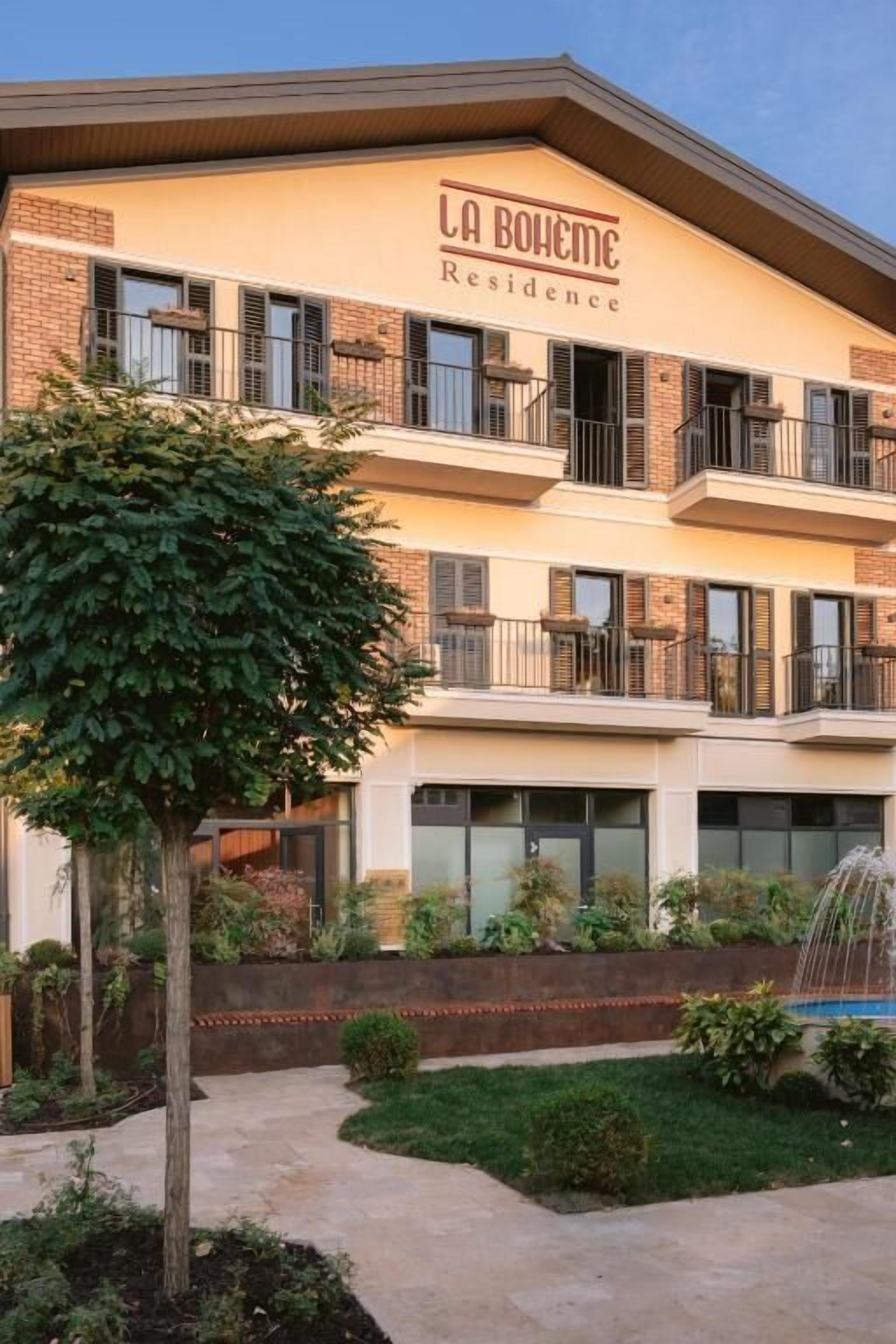 A multi-story building with balconies, a sign reading 'LA BOHEMIE Residence,' and a landscaped garden with a tree and a fountain.