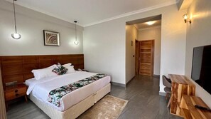 Un dormitorio con cabecera de madera, cama con ropa de cama blanca, una mesita de noche, un televisor y una mesita auxiliar de madera.