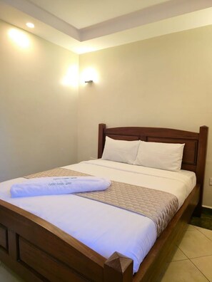 Una cama bien tendida con cabecera de madera, ropa blanca y una toalla doblada al pie.