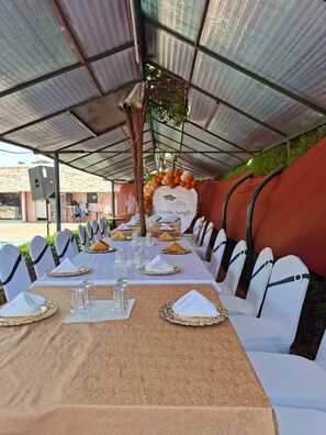 Un área de comedor al aire libre cubierta con mesas dispuestas para una comida, sillas con fundas blancas y un banner con un nombre.