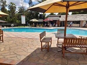 A swimming pool area with tiled flooring, wooden benches, and umbrellas providing shade. - Castle Gardens Hotel (Nairobi)