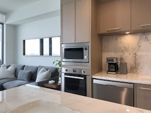 A modern kitchen with built-in appliances, a marble countertop, and a grey sofa in the adjacent living area. - Aqua Village - Pool, Sauna, Gym, Golf Sim (Kelowna)
