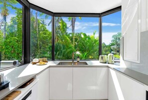 A modern kitchen with a large window, white cabinetry, and a view of lush greenery. - Belle Escapes 22 Rydges Plaza Esplanade with Garden Views (Cairns)