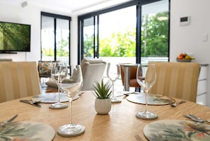 A dining table set with plates, cutlery, and wine glasses, with a small plant centerpiece, in a room with large windows and a television on the wall. - 22 Rydges Plaza Esplanade with Garden Views (Cairns)
